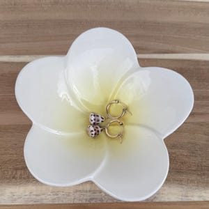 May include: A white ceramic flower-shaped dish with a yellow center holds a pair of gold hoop earrings with seashell charms. The earrings have small, speckled shells. The dish sits on a wooden surface.
