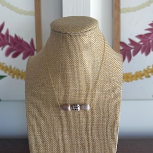 May include: A gold chain necklace with three decorative beads. The beads include a light pink, a black and white patterned, and a white bead. The necklace is displayed on a tan jewelry bust.