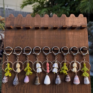 May include: A wooden display stand holds several keychains with silver rings. Each keychain features a unique seashell or decorative element, including white, pink, and green accents. The stand is made of dark brown wood.