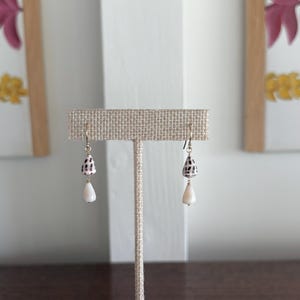 May include: A pair of dangle earrings with a seashell theme. Each earring features a small, speckled shell and a teardrop-shaped white bead. The earrings are suspended from gold-colored hooks and displayed on a neutral-toned stand.