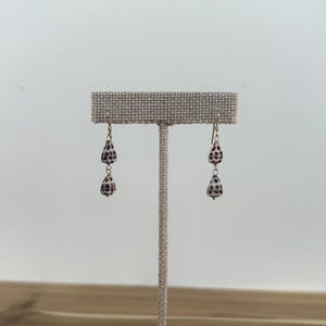 Puede incluir: Pendientes colgantes con cuentas en forma de lágrima con un patrón moteado en tonos marrón y blanco. Los pendientes cuelgan de ganchos dorados y se exhiben en un expositor de joyería de tonos neutros.