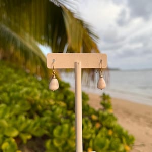 Puede incluir: Un par de pendientes en forma de lágrima, de color rosa pálido, expuestos en un soporte blanco. Los pendientes tienen ganchos dorados y pequeños detalles dorados. El fondo presenta una escena de playa con follaje verde y un cielo nublado.