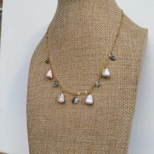 May include: A gold-colored necklace with white and light pink teardrop-shaped pendants and small green and gray stone accents. The necklace is displayed on a tan jewelry bust.