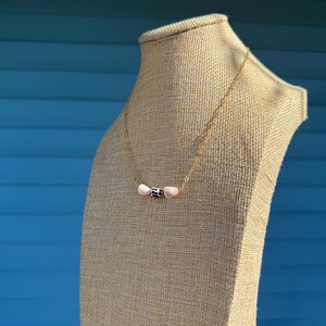 May include: A delicate gold necklace featuring a central pendant with a black and white patterned bead flanked by two light pink beads. The necklace is displayed on a tan mannequin against a blue background.