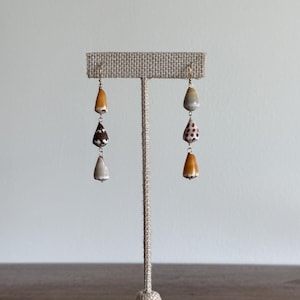Può includere: Orecchini con tre ciondoli a forma di conchiglia nei toni del marrone, beige e bianco. Gli orecchini sono esposti su un espositore per gioielli beige. Le conchiglie hanno motivi e texture uniche, con un gancio color oro.