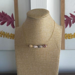 May include: A gold-colored chain necklace with four decorative beads. The beads are beige, white with black spots, clear, and red with white spots. The necklace is displayed on a tan jewelry bust.