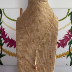 May include: A gold-colored necklace with a delicate chain and three pendant stones. The stones are white, off-white, and peach-colored, suspended in a vertical line. The necklace is displayed on a tan jewelry bust, with floral paintings in the background.