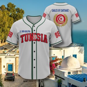K&ouml;nnte beinhalten: Wei&szlig;es Baseball-Trikot mit gr&uuml;nen Zierleisten, dem Wort "TUNISIA" in Rot und der Nummer 88 auf den &Auml;rmeln. Auf der R&uuml;ckseite des Trikots steht "EAGLES OF CARTHAGE" und ein kreisf&ouml;rmiges Emblem.