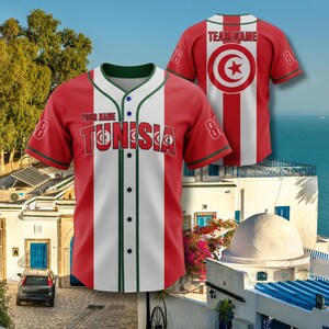K&ouml;nnte beinhalten: Rotes und wei&szlig;es Baseball-Trikot mit dem Wort "TUNISIA" auf der Brust. Das Trikot hat gr&uuml;ne Verzierungen und die Nummer 8 auf den &Auml;rmeln. Die R&uuml;ckseite des Trikots ist rot mit einem wei&szlig;en Streifen und einem kreisf&ouml;rmigen Emblem mit einem Stern.