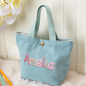 May include: A light blue corduroy tote bag with a matching handle and the name "Amelia" embroidered in pink and peach. The bag has a button closure and is sitting on a white fluffy surface. Pens are visible in the lower left corner.