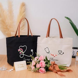 May include: Two tote bags, one black and one off-white, with brown leather handles. Each bag features a stethoscope design with floral accents and a name. A bouquet of pink roses and a card are in front of the bags.