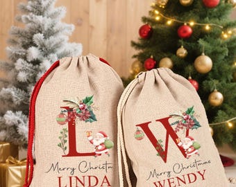 Puede incluir: Dos bolsas de regalo navideñas beige con cordón. Una bolsa presenta la letra roja "L" con adornos florales, una ilustración de Papá Noel y el texto "Merry Christmas LINDA". La otra bolsa tiene una "W" roja y el texto "Merry Christmas WENDY".