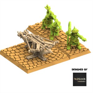Op de afbeelding: Een miniatuurmodelset met een lichtgekleurde houten katapult en drie groene krijgersfiguren. De figuren staan op bruinachtige, getextureerde platforms. De afbeelding bevat de tekst "DESIGNED BY" en een logo.