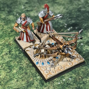 Könnte beinhalten: Miniaturfiguren von zwei Kriegern mit einer Armbrust. Eine Figur hält ein Buch, die andere einen Speer. Sie tragen silberne Rüstungen mit roten und weißen Akzenten. Die Armbrust ist mit goldenen und blauen Elementen detailliert, auf einer beigen Basis.