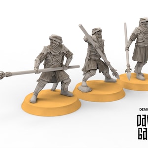 Może przedstawiać: Trzy szare miniaturowe figurki wojowników w zbrojach, z których każdy trzyma włócznię. Figurki stoją na okrągłych, beżowych podstawach. Tekst "DESIGNED BY DAVALE GAMES" znajduje się w prawym dolnym rogu.