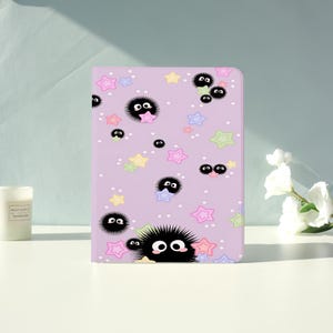 May include: A lavender-colored notebook featuring a repeating pattern of black, fluffy cartoon characters with large eyes, and colorful stars. The notebook is on a white surface with a candle and flowers in the background.