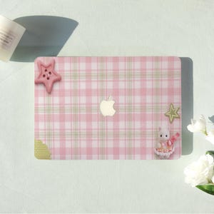 May include: A pink and white plaid laptop case with a white Apple logo. The case is decorated with a pink star-shaped button, a green star, and a small figurine of a rabbit wearing a dress. The case is on a light green surface.
