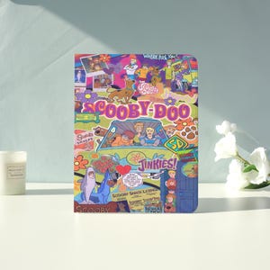 May include: A colorful Scooby-Doo themed notebook featuring the Mystery Machine van and the gang. The cover includes the text "WHERE ARE YOU!" and "JINKIES!" along with various characters and the Scooby-Doo logo. The background is a mix of floral and cartoon elements.
