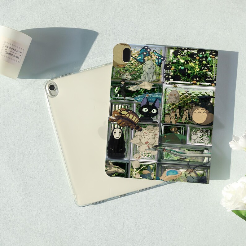 May include: A white tablet with a colorful case featuring various anime characters. The case has a grid-like design with images of a black cat, a turtle, and other characters. A white candle is in the background.