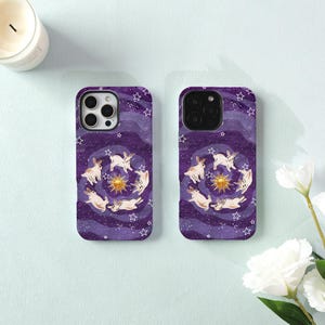 May include: Two smartphone cases featuring a deep purple background with a swirling galaxy pattern, white stars, a central golden sun, and multiple white rabbits arranged in a circular formation around the sun on each case.