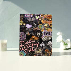 May include: A rectangular notebook featuring a collage of Halloween-themed stickers, including pumpkins, ghosts, witches, and skeletons. The cover displays the text "Doin' Witchy Shit" in large pink letters, alongside "Let's Get Spooky" and "All Hallows Eve."