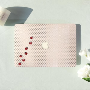 Peut inclure: Un autocollant pour ordinateur portable avec un fond crème clair, un motif répétitif de petits points noirs et une traînée de sept coccinelles rouges à points noirs disposées en ligne courbe à gauche du logo Apple argenté central.