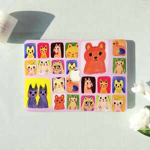 Op de afbeelding: Een rechthoekige laptopskin met een lichtroze achtergrond bedekt met een raster van kleurrijke, handgetekende illustraties van diverse katten en kleine dieren in gele, oranje, paarse en groene kaders met een Apple-logo uitsparing.