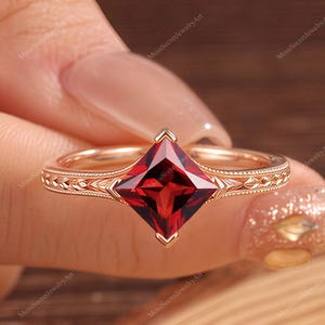 Può includere: Un anello in oro rosa con una pietra preziosa granato rosso scuro taglio princess montata su quattro griffe. La fascia presenta intricati dettagli incisi sui lati, conferendo un aspetto strutturato alla superficie del metallo.