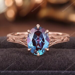 Può includere: Un anello in oro rosa con una pietra preziosa dal taglio ovale che cambia colore tra tonalità blu e viola. La fascia presenta incisioni floreali dettagliate lungo i lati e la pietra è fissata da sei griffe in una montatura classica.