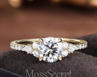 Può includere: Un anello in oro con una grande pietra centrale rotonda taglio brillante, affiancata da pietre d'accento più piccole rotonde e a forma di pera su una fascia sottile. Il testo MossSecret JewelryArt è visibile sotto l'anello. Montatura a griffe.