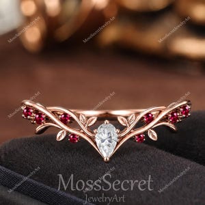 Può includere: Un anello in oro rosa con una pietra centrale trasparente a forma di pera, affiancata da piccole pietre rosse incastonate in una fascia a forma di ramo con dettagli a foglia. L'anello è in una scatola scura con il testo MossSecret JewelryArt.