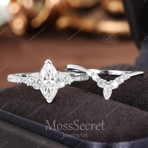Pode incluir: Um conjunto de anéis de noivado em tom prateado com uma pedra central de corte marquise ladeada por pedras redondas e marquise, acompanhado por uma aliança em forma de V adornada com uma série de pedras redondas e uma pedra de corte marquise.