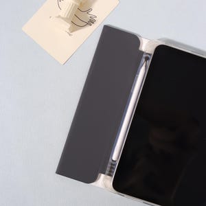 May include: A black tablet case with a clear frame, holding a tablet and a white stylus. The case is open, revealing the tablet's black screen. A small white sculpture and a card with a drawing are in the background.