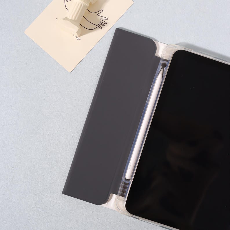 May include: A black tablet case with a clear frame, holding a tablet and a white stylus. The case is open, revealing the tablet's black screen. A small white sculpture and a card with a drawing are in the background.