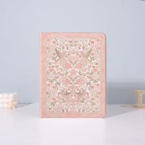 May include: A pink journal with a floral and bird pattern. The cover features a symmetrical design with pink flowers, green leaves, and birds. The edges of the journal are also pink, creating a cohesive look. The journal is rectangular and appears to be closed.