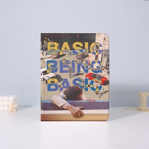 Könnte beinhalten: Ein rechteckiges Notizbuch mit einem Collage-Design. Das Cover zeigt die Worte "BASIC BEING BASIC" in Gelb und Blau, mit einem Hintergrund aus Autos und Menschen. Eine Person lehnt über einer Brüstung.