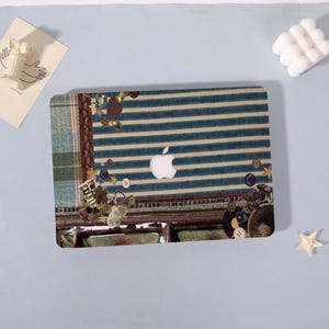 May include: A laptop case featuring a blue and cream striped pattern with a white Apple logo. The case is decorated with various buttons, stars, and fabric patches. The design incorporates a plaid section and a brown border.