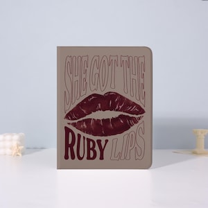 May include: A rectangular notebook with a taupe cover featuring a graphic of ruby red lips and the text "SHE GOT THE RUBY LIPS" in a matching colour. The design is centered on the cover.