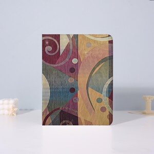 May include: A rectangular notebook featuring a vibrant abstract pattern with overlapping geometric shapes, curved lines, and circles in shades of deep burgundy, mustard yellow, teal, and muted pink, all set against a fine vertical line texture.