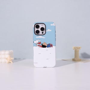 May include: A white and blue phone case featuring a cartoon illustration of Snoopy and Charlie Brown. Snoopy is wearing a red cape and lying on a cloud with Charlie Brown. The case has a blue sky background with white clouds.