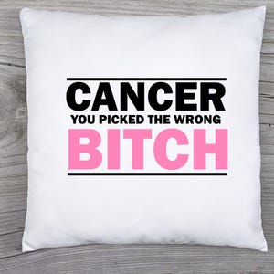 May include: A white square pillow with the text "CANCER YOU PICKED THE WRONG BITCH" in black and pink. The text is centered on the pillow, with the word "CANCER" at the top and "BITCH" at the bottom.