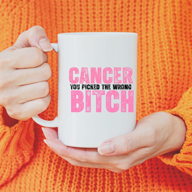 May include: White ceramic mug with the text "CANCER YOU PICKED THE WRONG BITCH" in pink and black lettering. The mug is held by a person wearing an orange sweater.