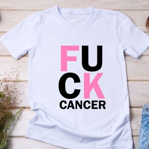 May include: White t-shirt with the words "FUCK CANCER" in a bold, blocky font. The letters "FU" and "C" are pink, while "U" and "K" are black. The word "CANCER" is in black.