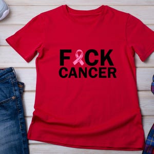 May include: Red t-shirt with the text "F*CK CANCER" in black, with the letter "C" replaced by a pink awareness ribbon. The shirt is short-sleeved and made of a soft material. The shirt is laying on a light wood surface.