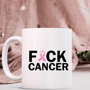 May include: White ceramic mug with a black and pink graphic. The mug features the words "F*CK CANCER" in a bold, black font, with the letter "C" replaced by a pink awareness ribbon. The mug has a white handle and is set against a soft, pink background.