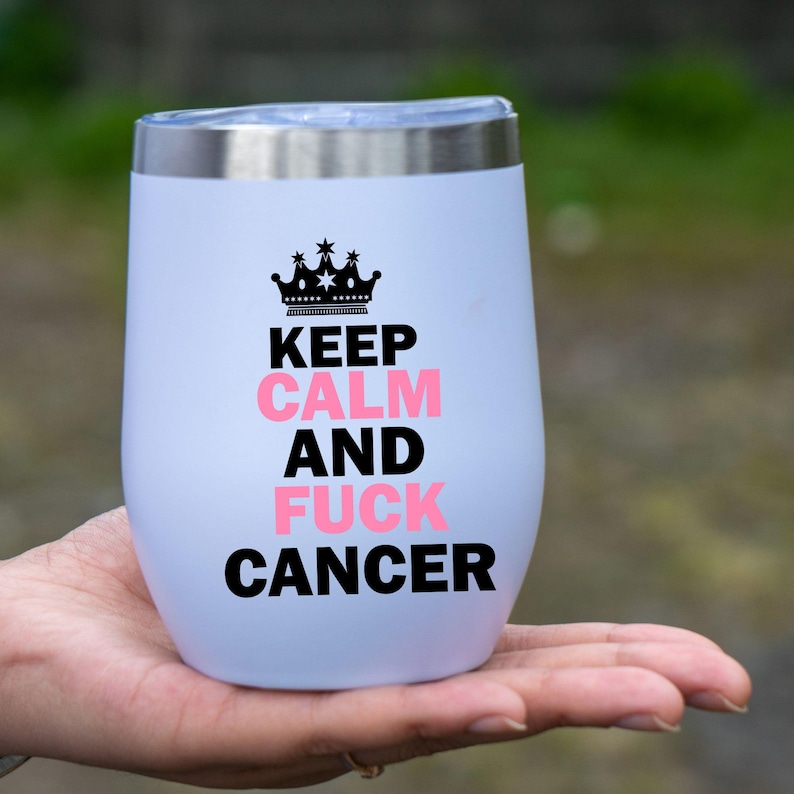 May include: A white, stemless wine tumbler with a silver rim. The tumbler features a black crown graphic above the words "KEEP CALM AND FUCK CANCER" in black and pink lettering. The tumbler is held in a person's hand.