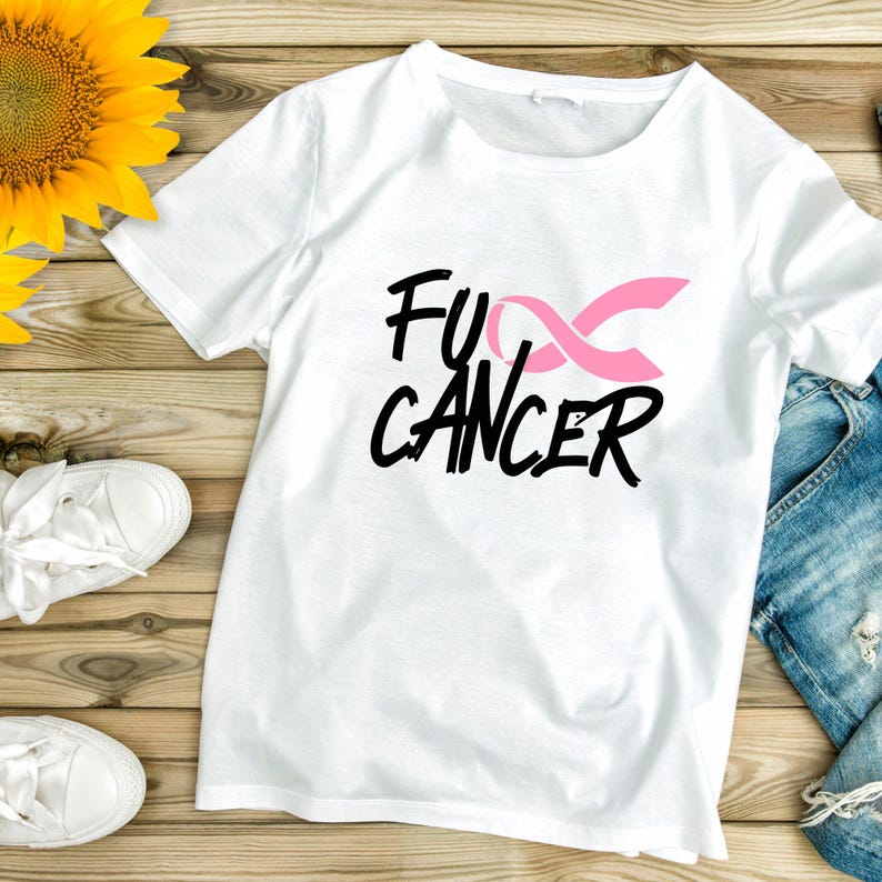 May include: White t-shirt with the text "Fu Cancer" in black script and a pink ribbon graphic. The shirt is displayed on a wooden surface with a sunflower and white sneakers.