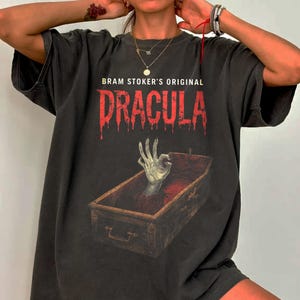 May include: Dark gray t-shirt featuring the text "Bram Stoker's Original Dracula" in dripping red letters. The graphic includes a hand emerging from a coffin. The shirt has a vintage, worn look.