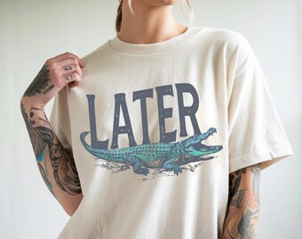 May include: Cream-colored t-shirt featuring the word "LATER" in dark blue block letters above a detailed illustration of a green and blue alligator with an open mouth. The shirt has short sleeves and a relaxed fit.