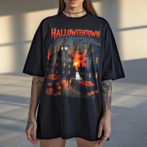 Puede incluir: Camiseta negra con un diseño de Halloween. El diseño muestra una escena de pueblo con calabazas, un fantasma y murciélagos, y el texto "Halloweentown" en naranja. La camiseta tiene un corte holgado.